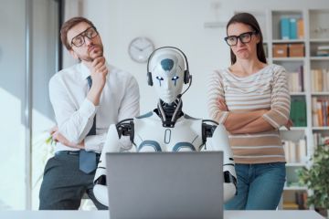 Humans look over the shoulder of a robot at a computer, symbolising human independent thought