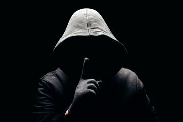 Shadowy figure with a hood holds a finger up to their face