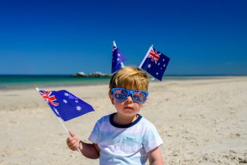 A young child on a beach wearing two Australia flags on his head waves another Australia flag