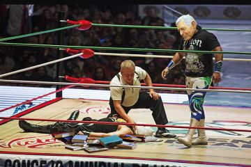 In a wrestling ring filled with books, one man lies prone and is being counted out while his masked opponent prowls