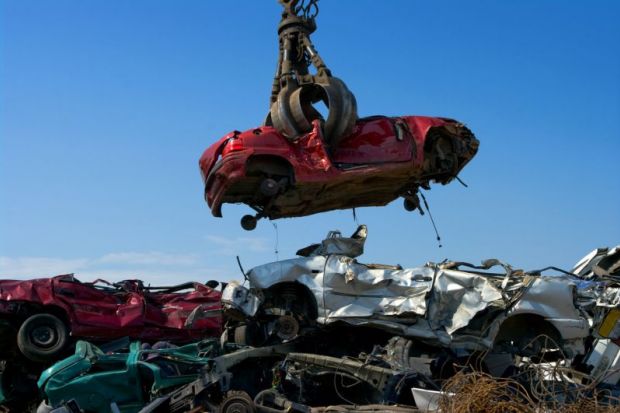 A shattered car is lowered into a pile of other crushed cars