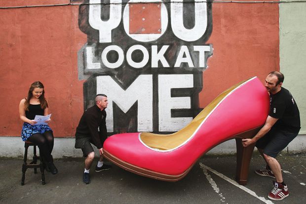 Two men carrying a big red shoe while a young woman sits reading