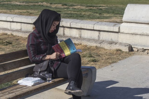 Isfahan, Iran - November 10, 2019. A young Iranian girl in modern clothes and a black shawl on her head reads a book on a bench in Imam Square.