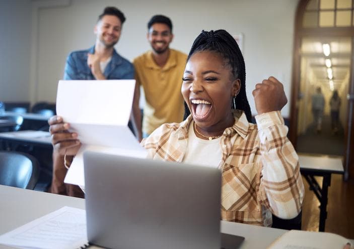 young woman celebrating receiving a scholarship award next to her computer with her two friends smiling in the background